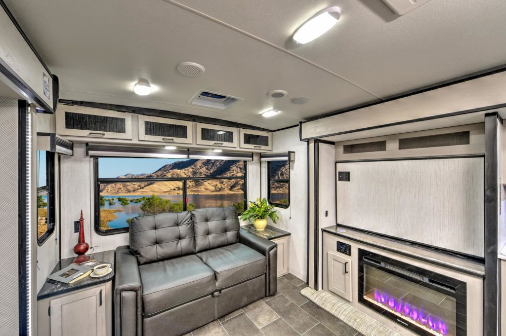 Modern RV interior with gray sofa, electric fireplace, and built-in cabinets. Large windows reveal scenic views of mountains and a lake. Natural light brightens the cozy, well-organized space with contemporary décor.