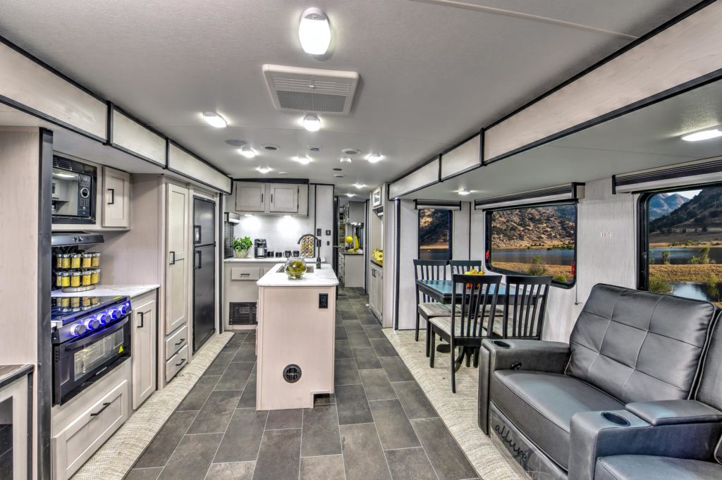 Modern RV interior with a kitchen on the left, featuring an oven, stove, refrigerator, and shelves, an island in the center, a dining table by large windows with mountain views, and a sofa on the right.
