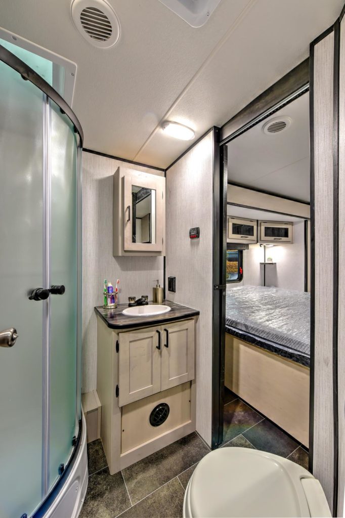 A compact RV bathroom with a rounded glass shower, small sink and cabinet, mirror, and toothbrushes; adjacent to a bedroom with a made bed and overhead storage.