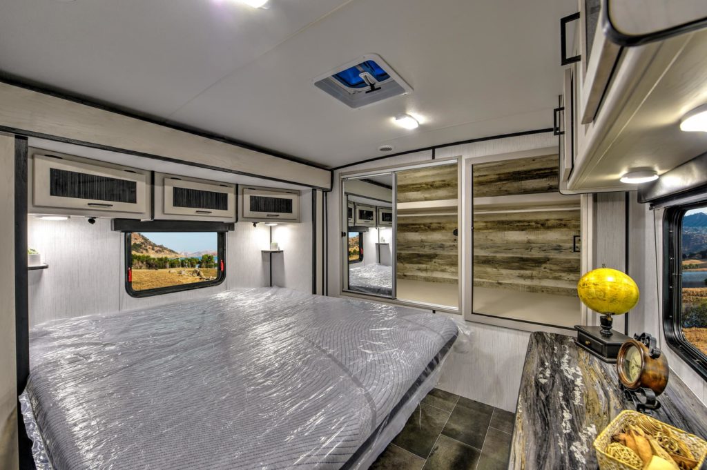 Interior of a modern RV bedroom featuring a large bed with plastic-wrapped mattress, overhead cabinets, a window with scenic views, built-in shelves, and a countertop with a decorative globe.