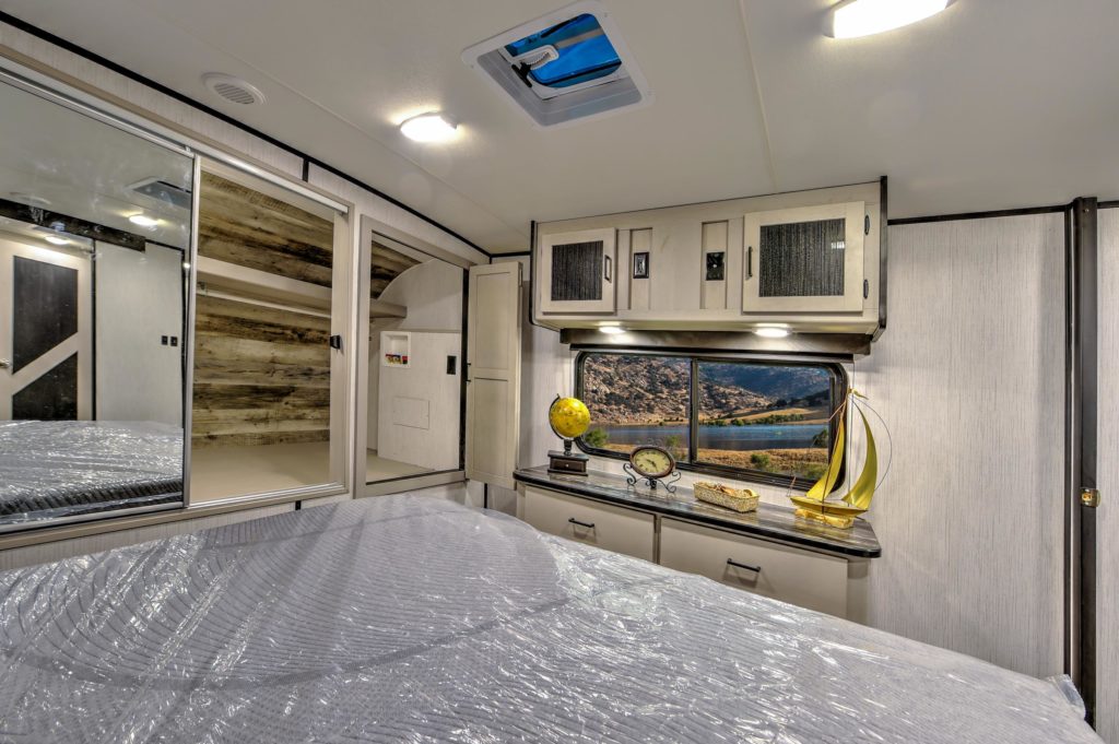Interior of a modern RV bedroom with a bed covered in plastic, built-in cabinets, mirrored closet doors, and a decorative shelf featuring a globe, a clock, a model sailboat, and a framed landscape photo.