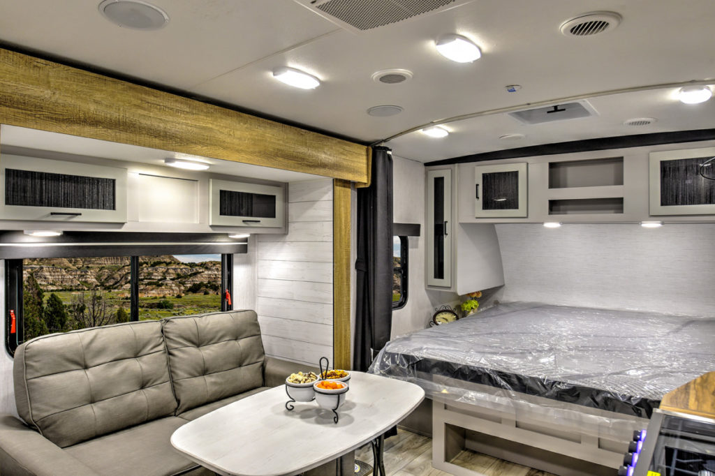 Interior of a modern RV featuring a gray sofa and dining table on the left. A bed with overhead storage is on the right. Large windows offer a view of a scenic landscape. The decor includes wood accents and bright lighting.