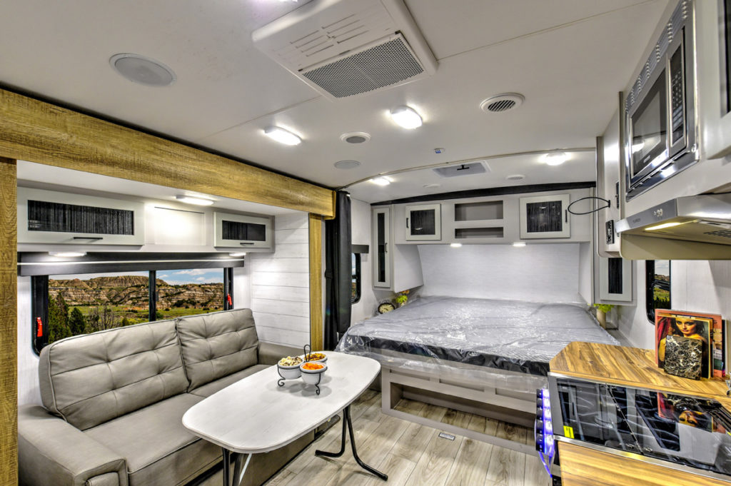 Interior of a modern RV featuring a fold-out bed, a dining area with a gray sofa and table, and a kitchenette equipped with a microwave and stove. The windows provide a scenic view of a rocky landscape outside.