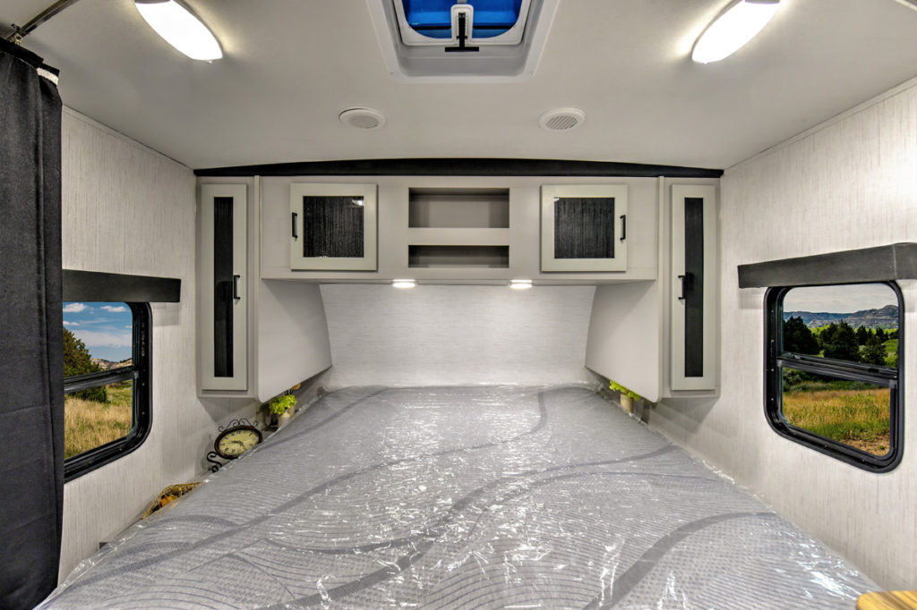 Interior of a camper trailer bedroom with a double bed covered in plastic. Two small windows are on either side, and there are cabinets above the head of the bed. The room is well-lit, and the windows reveal a view of grass and trees outside.