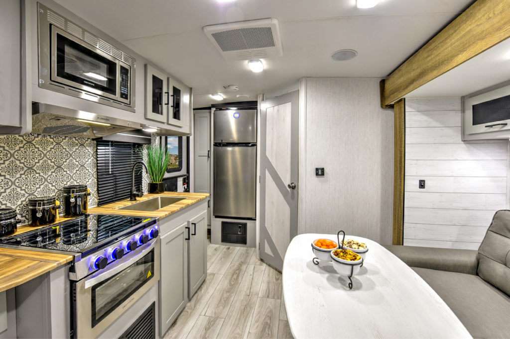 Modern RV interior featuring a compact kitchen with stainless steel appliances, tiled backsplash, and wooden countertops. A small dining table holds a fruit bowl. Gray seating and light wood accents complete the cozy space.