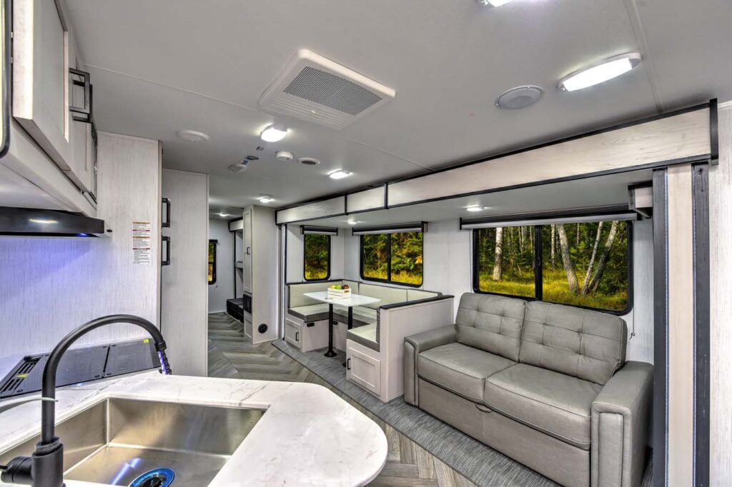 Modern RV interior with light wood cabinets, a gray pull-out sofa, a built-in dining area with benches and table, large windows showing trees, and a marble-look sink in the foreground.