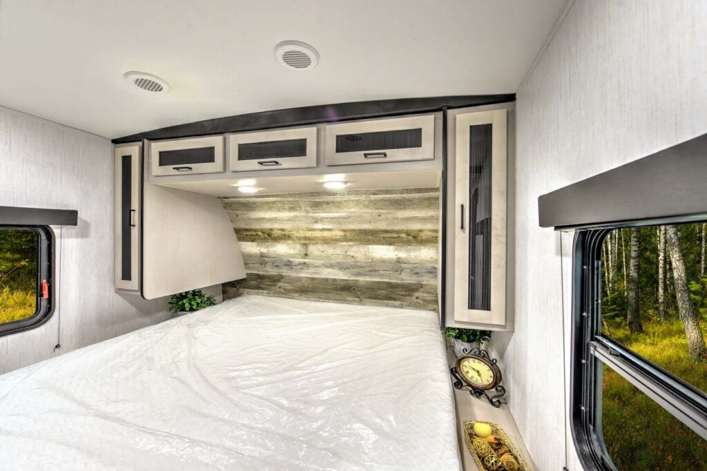 A neatly made bed with a white cover inside an RV, featuring overhead cabinets, reading lights, a small clock, and a window showing a forested outdoor scene.