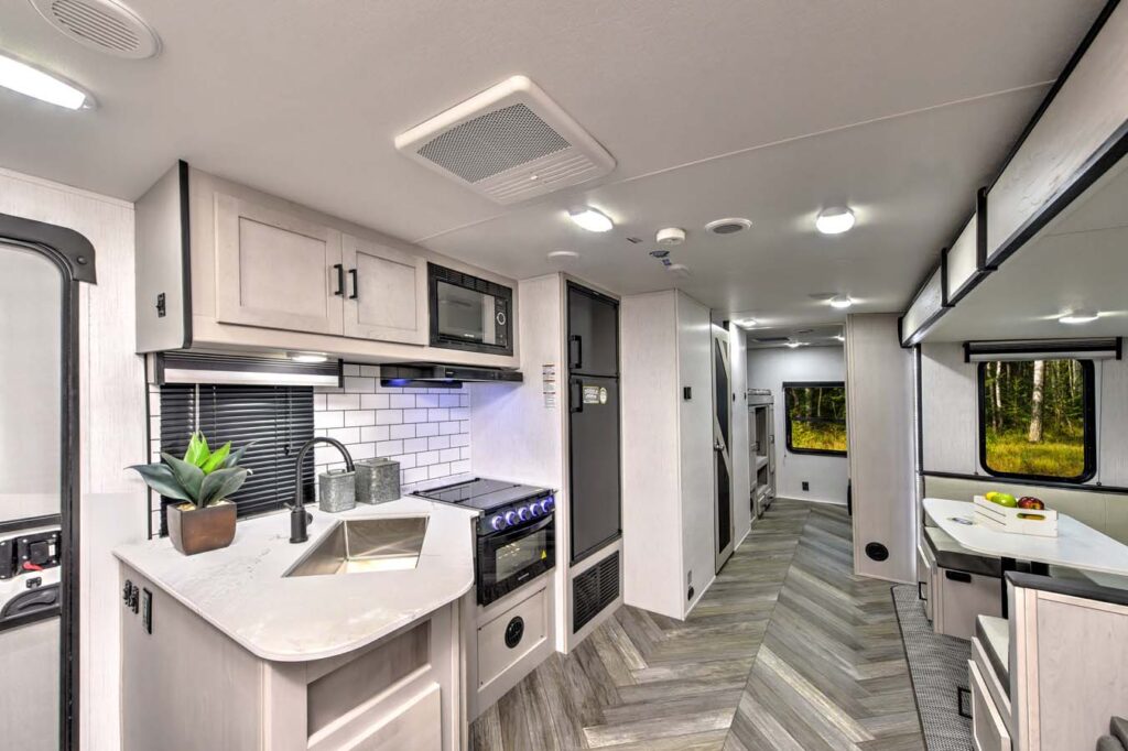 Modern RV interior with a kitchen featuring white cabinets, a sink, stove, oven, microwave, and subway tile backsplash. A dining area and hallway with wood-patterned flooring are visible, with windows showing trees outside.