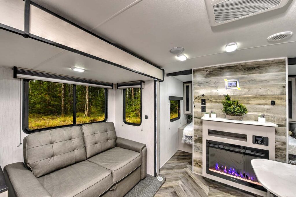 Modern RV interior with a gray sofa, large windows showing a forest view, and a cozy area featuring an electric fireplace, potted plants, and a bed partially visible in the background.