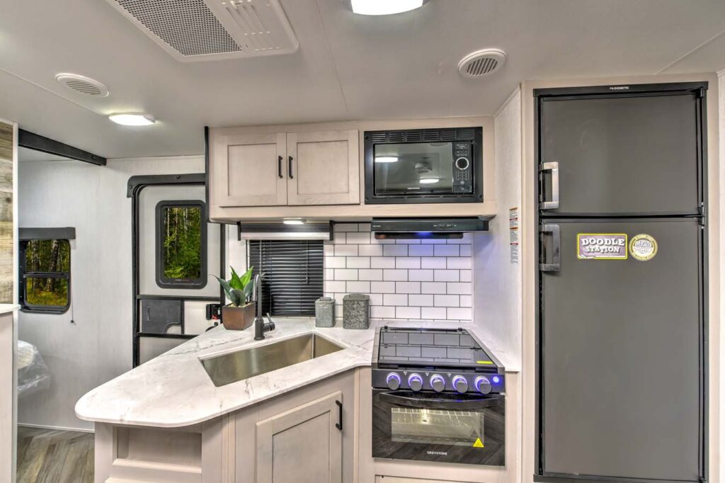 Modern RV kitchen with a marble-patterned countertop, sink, stove, oven, microwave, small potted plant, and a refrigerator with a “Doodle Station” sticker. Subway tile backsplash and forest view through the window.