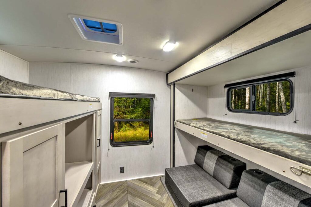 Interior of an RV with bunk beds, a window showing a forest view, a cushioned seating area below one bunk, wood-patterned flooring, and overhead lighting. The decor is modern and neutral-toned.
