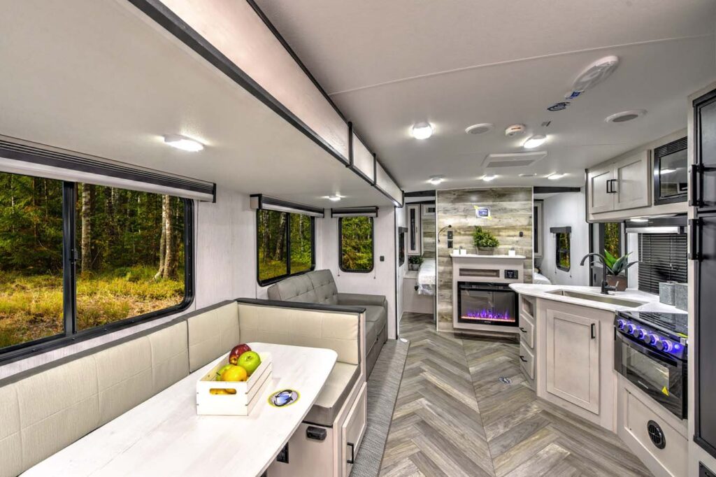 Interior of a modern RV with a dining booth, sofa, fireplace, TV, kitchen area, and large windows showing a forest outside. Neutral colors and wood accents create a cozy, stylish atmosphere.