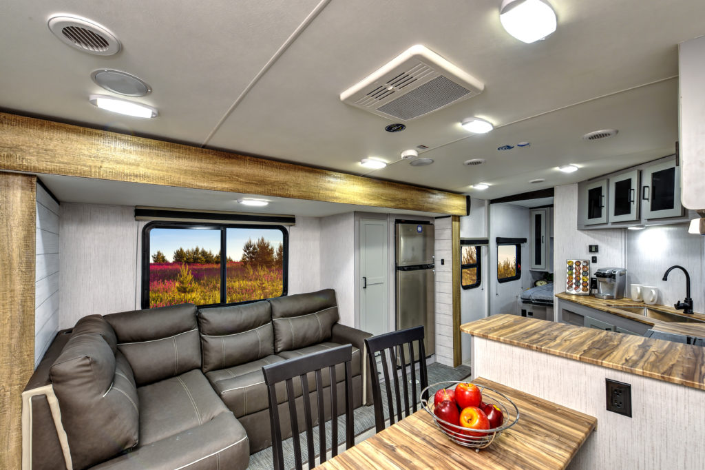 Interior of a camper van with a modern living space, featuring a gray L-shaped sofa, dining table with chairs, and a kitchenette. Large windows show an outdoor view of colorful wildflowers and trees. A bowl of apples sits on the counter.