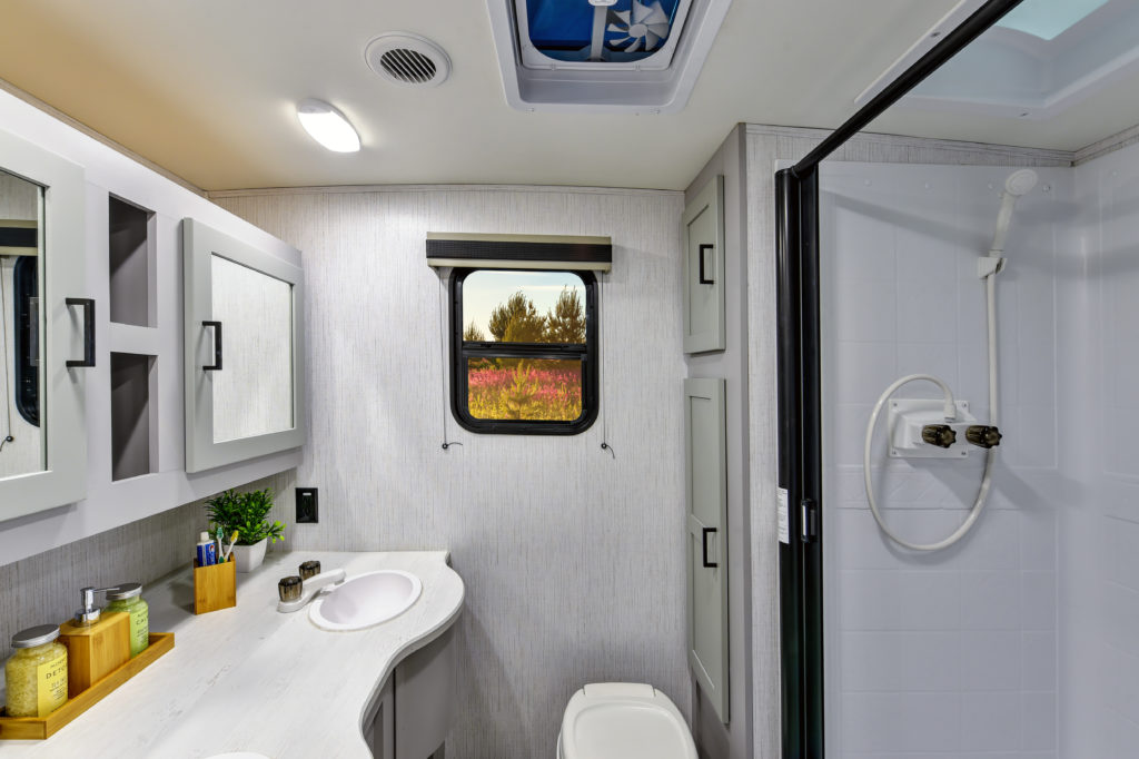 Modern RV bathroom with white cabinets, a curved sink, and a toilet. A shower with a glass door is on the right, and a small window above the sink shows an outdoor scene with trees and flowers. Toiletries sit on the countertop.