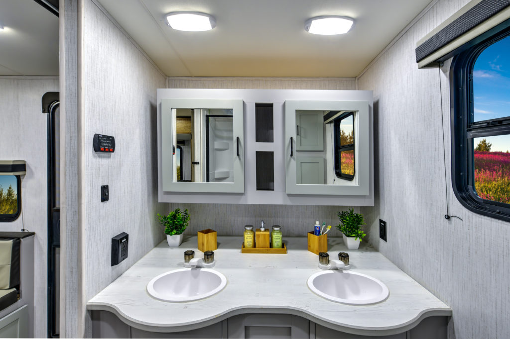 Modern RV bathroom with two sinks, a white countertop, and mirrored cabinets. The counter has green plants, soap dispensers, and amenities. A window shows a scenic outdoor view with flowers and trees under a blue sky.