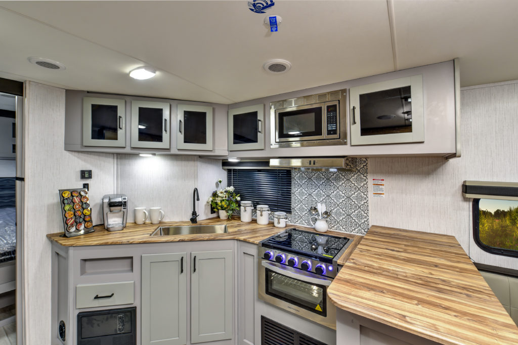 A modern RV kitchen with wooden countertops, gray cabinets, and a stove. Theres a backsplash with a patterned design, a microwave, a coffee maker, and assorted kitchenware, including mugs and canisters on the counter.