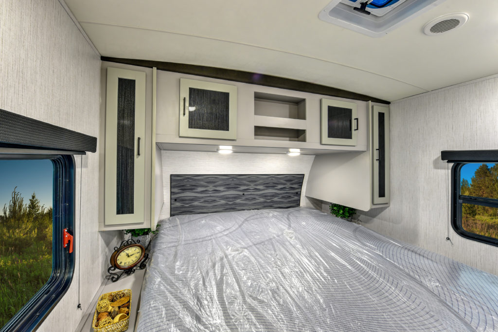 Interior of a camper bedroom featuring a bed with plastic covering, overhead cabinets, small plants, a vintage-style clock, and a window offering a view of greenery outside. Soft lighting creates a cozy atmosphere.