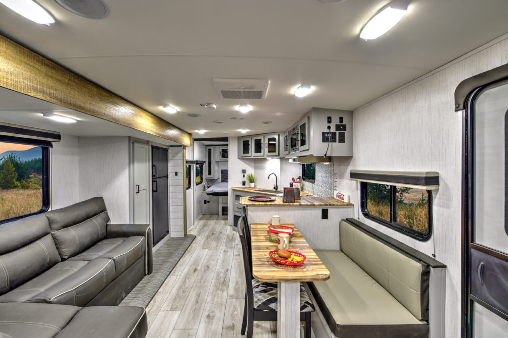 Interior of a modern RV featuring a gray sofa, wooden dining table with place settings, kitchen area with sink and appliances, and large windows. Light wood flooring and white walls create a spacious and well-lit atmosphere.