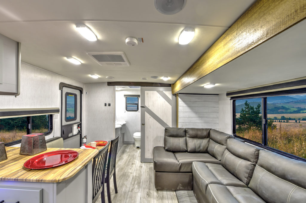 Interior of a modern RV with a cozy living area featuring a leather sectional sofa, a dining table with red plates, and a kitchen counter. Large windows offer a scenic view of fields and hills. Bright ceiling lights illuminate the space.