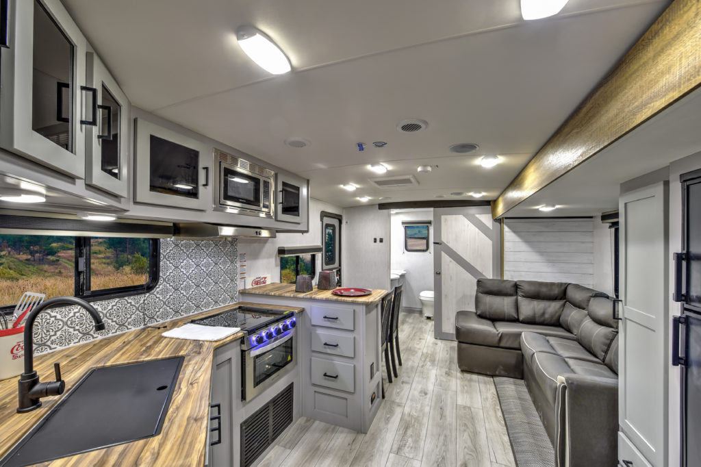Interior of a modern RV with a kitchen on the left featuring a sink, stovetop, and microwave. On the right, theres a gray leather sofa. The design is sleek with wooden countertops and light gray cabinetry.