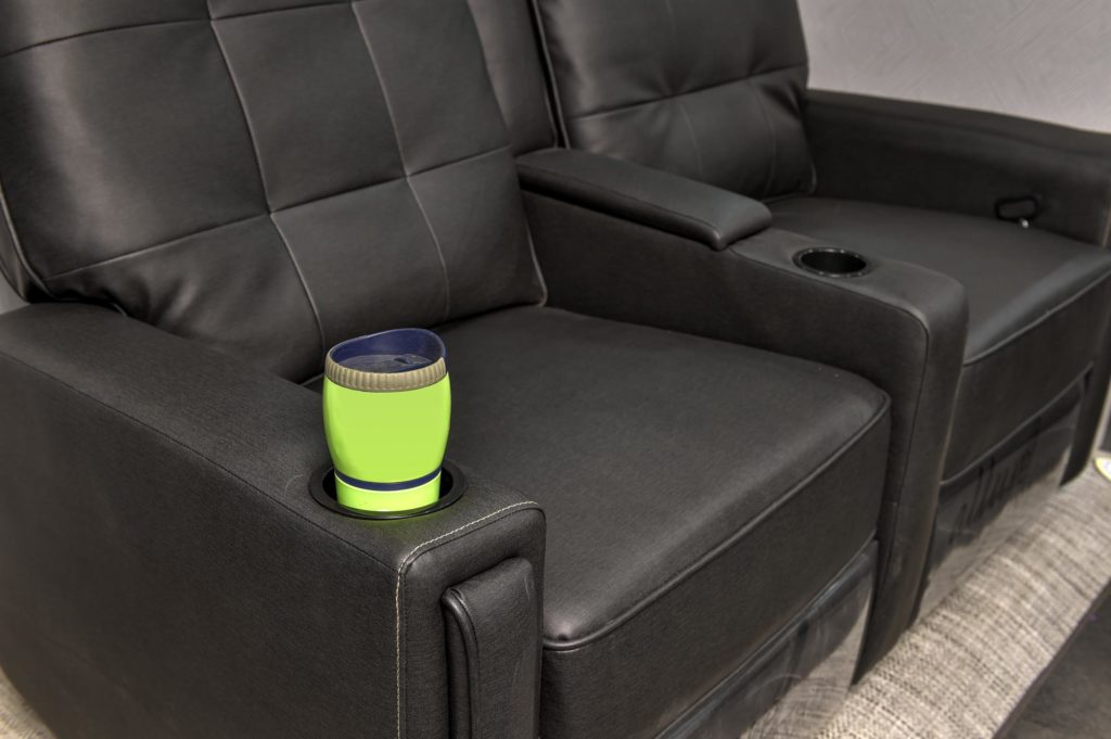 A bright green cup with a dark lid sits in the cup holder of a black leather recliner sofa, next to another empty seat. The sofa is on a gray carpet and has a modern design.