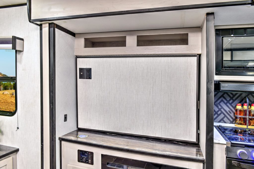 A built-in TV lift cabinet in a modern RV interior, with the TV hidden behind a textured panel above a wood-finished shelf, next to a kitchen area with a stove and spice bottles.