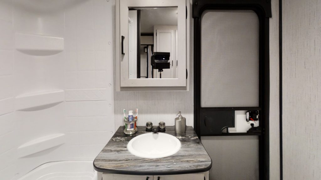 A compact bathroom with a sink, two toothbrushes, soap dispenser, mirror cabinet, and a black-framed frosted door. A white shower with built-in shelves is partially visible on the left.