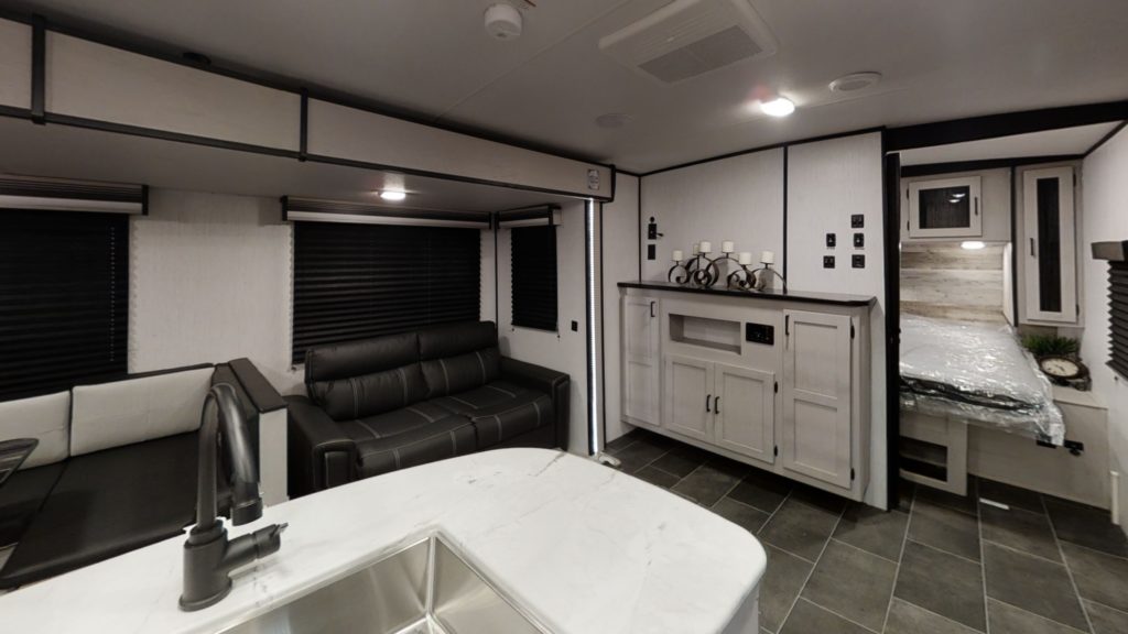 Interior of a modern RV with a kitchen sink in the foreground, a black sofa, a dining booth, cabinetry on the right, and a partially visible bed in a separate sleeping area toward the back.