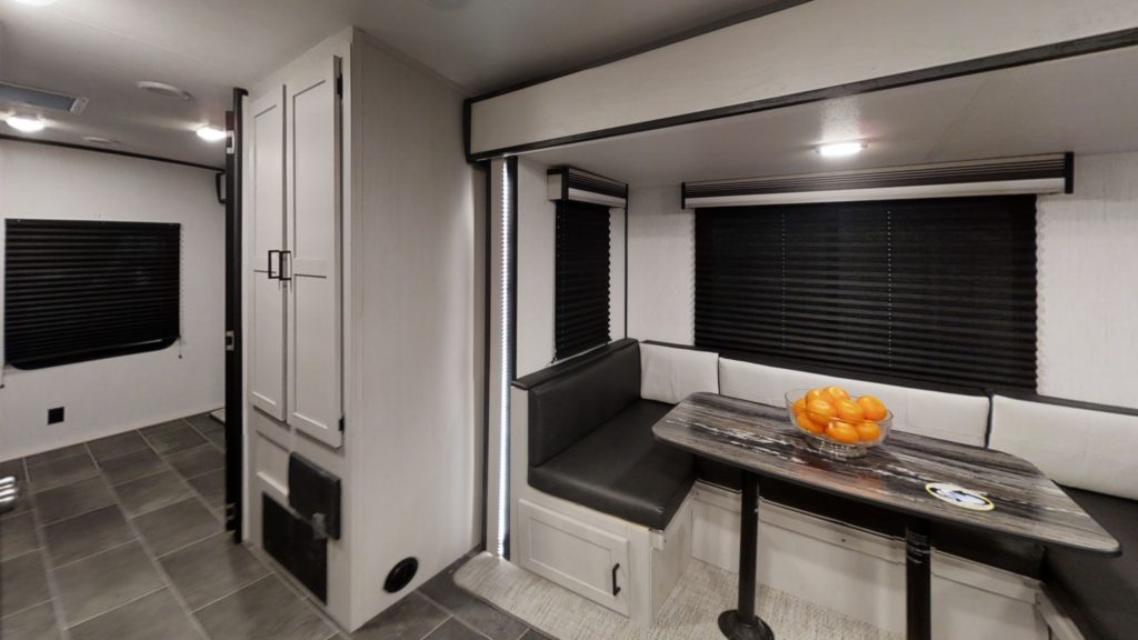 A modern RV interior with gray cabinets, black-out blinds, and a dining area featuring a black cushioned bench, a table with a woodgrain finish, and a bowl of oranges on top. The flooring is dark tile.