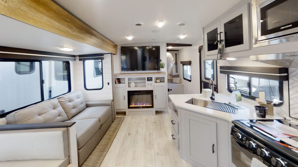 The interior of this modern, compact living space is reminiscent of ultra-lightweight travel trailers, showcasing a kitchen with stove and sink, a comfortable sofa, large windows, and a wall-mounted TV above the fireplace. Light wooden flooring and décor create an inviting atmosphere.