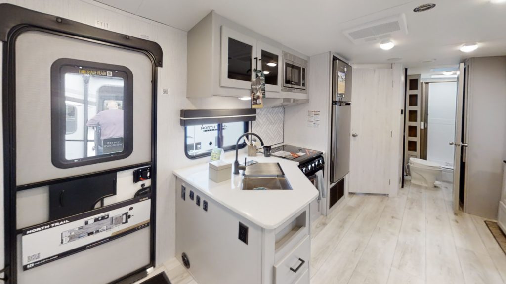 The interior of this ultra-lightweight travel trailer features a compact kitchen with a stainless steel sink, stove, and overhead microwave. Light wood cabinetry and flooring enhance the spacious feel. A doorway leads to a bathroom with a visible toilet, next to the exterior door.