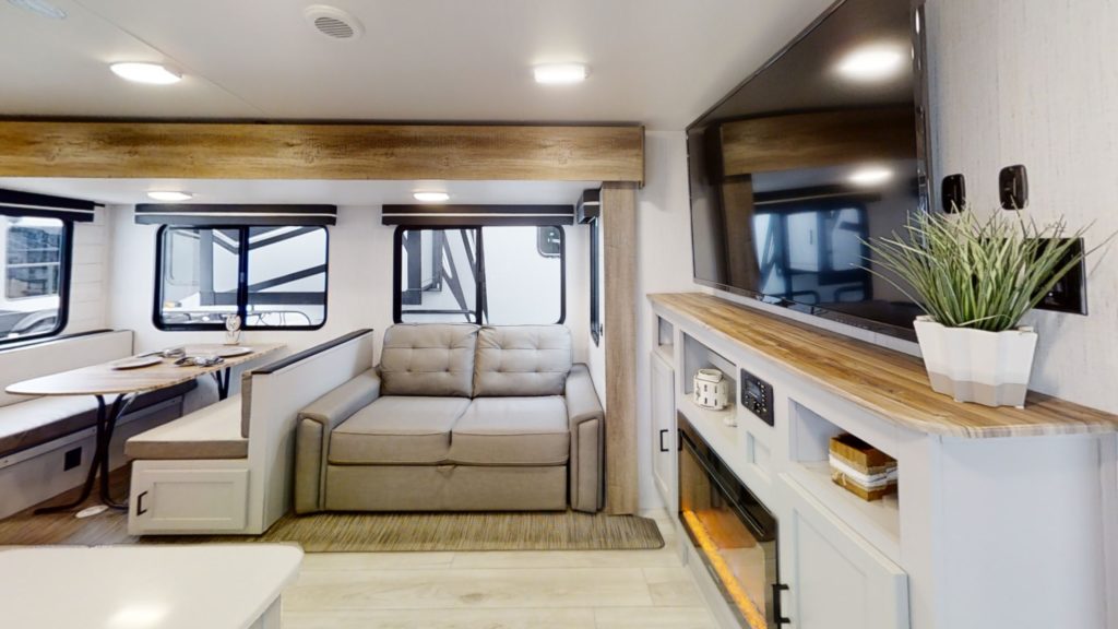 The interior of this ultra-lightweight travel trailer features a modern RV living area with a grey sofa, wall-mounted flat-screen TV, and decorative plant on a wooden shelf. On the left, youll find a cozy dining area with a circular table and window.