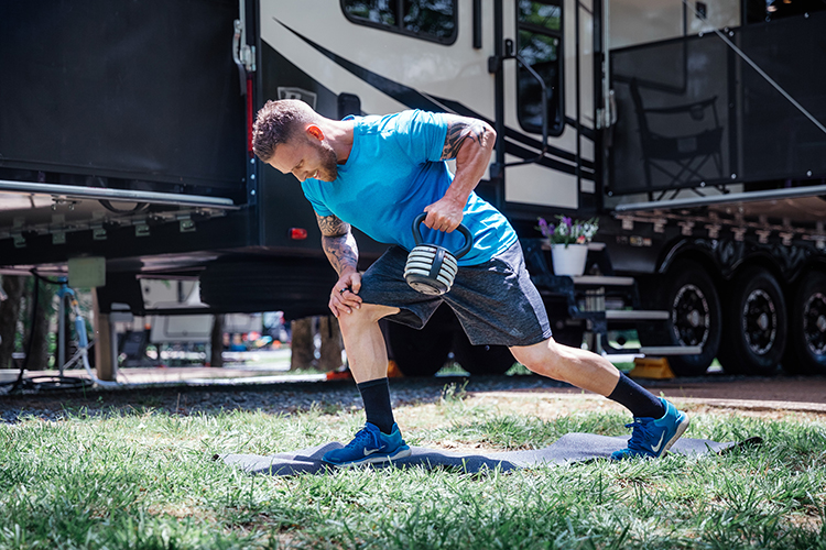 A fit man doing one-armed rows on a yoga mat outside his RV at a camp site.