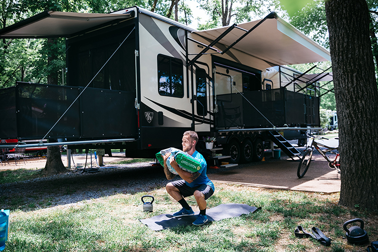 A man holds a big bag of dog food as a weight while he does squats outside his RV.