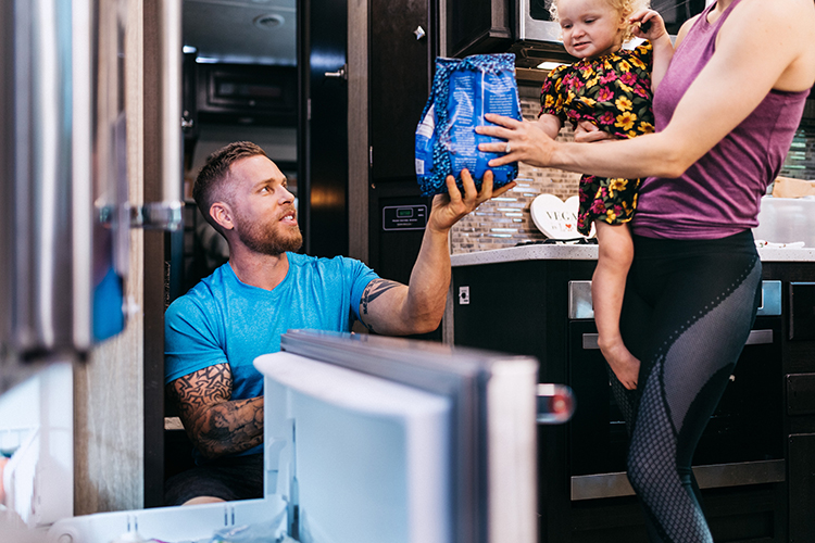 A man knelt in front of a freezer drawer, handing a woman a bag of frozen blueberries.