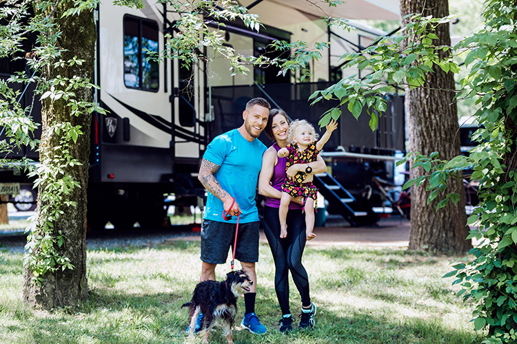 A young family holding a baby and standding with a dog on a leash, standing outside their RV at a campsite and smiling.
