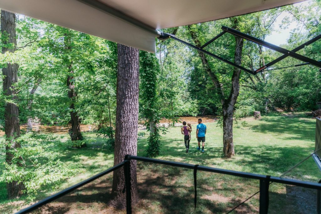 Two adults and a young child seen from the patio of a toy hauler RV, walking through trees toward a stream.