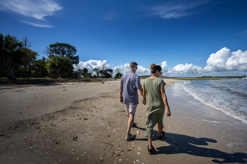A couple walking down the beach holding hands, with an RV in the distance on the shore.