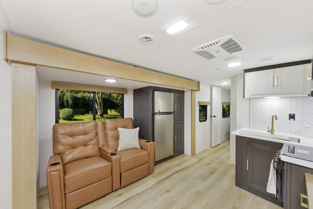 Interior of a modern RV featuring two brown leather recliners with a cushion, a compact kitchen with white cabinets and a chevron backsplash, a stainless steel refrigerator, light wood flooring, and large windows with views of trees outside.