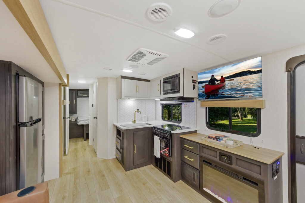 Modern RV interior with wood flooring, a compact kitchen featuring a stove, microwave, and large fridge. A countertop area includes a TV showing a canoe scene. Through a doorway, a bed is visible, suggesting a sleeping area. Natural light enters from windows.