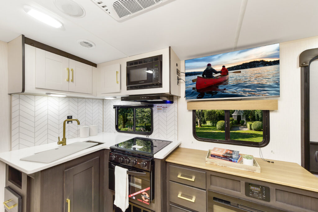 Interior of an RV kitchen with modern appliances and white cabinets. A flat-screen TV displays two people in a red canoe on a lake. A window offers a view of a green park.