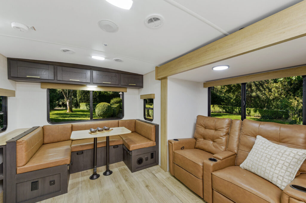 Interior of an RV featuring a light wood floor and brown leather furnishings. The space includes a U-shaped booth with a table, overhead cabinets, and two reclining chairs with a pillow. Large windows offer views of greenery outside.