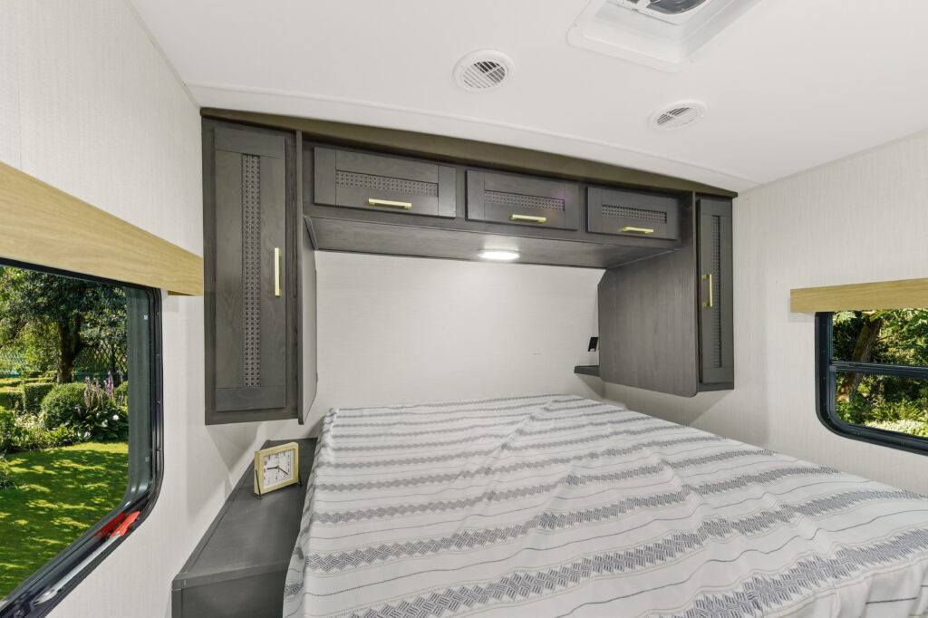 Interior of a recreational vehicle bedroom with a neatly made bed featuring a patterned duvet. The room has dark wooden cabinets above the bed, a small clock on a shelf, and windows showing greenery outside.
