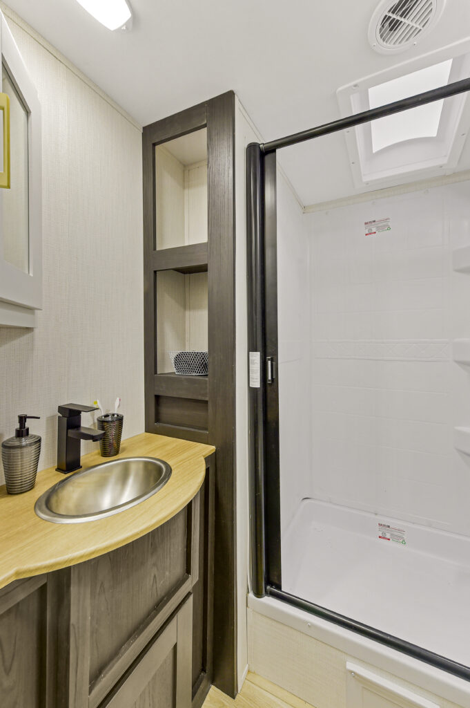 Modern bathroom with a wooden vanity, stainless steel sink, and black faucet. Adjacent to the vanity is a spacious glass-door shower. Above the sink is a shelf with decorative items. Neutral color palette and a ceiling light provide a bright atmosphere.