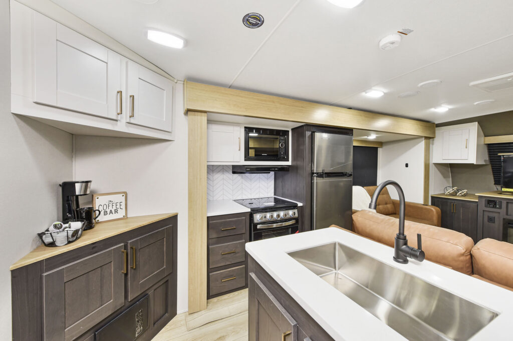 Modern RV kitchen interior with a stainless steel sink, black faucet, stovetop, oven, microwave, refrigerator, coffee maker, and Coffee Bar sign next to brown leather couches and light cabinets.