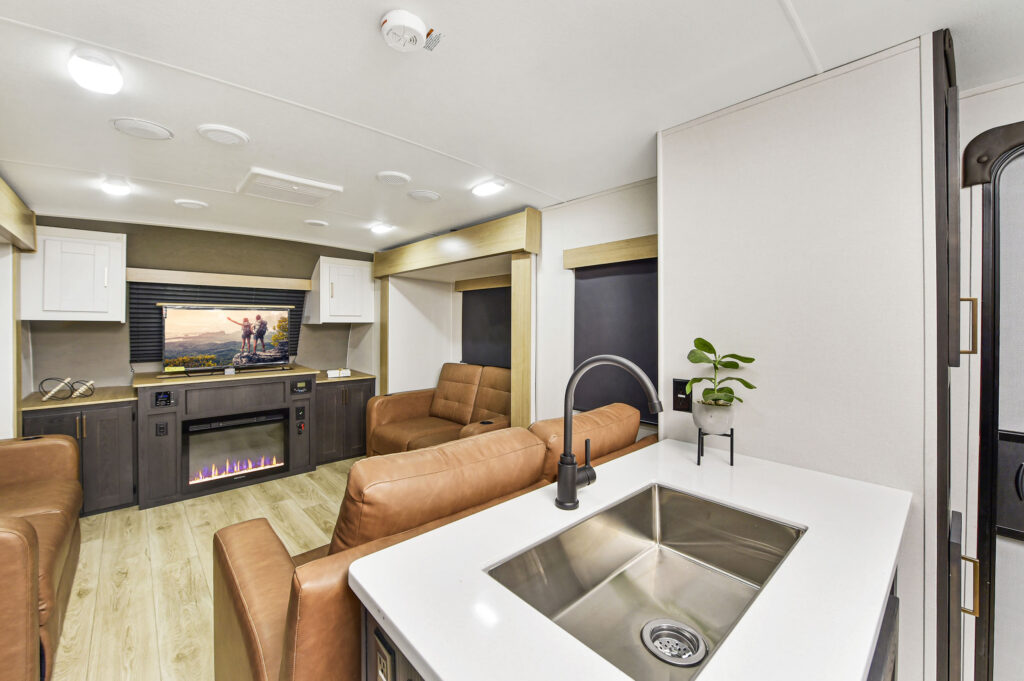 Modern RV interior with brown leather sofas, a TV above an electric fireplace, wood flooring, and a kitchen area featuring a stainless steel sink, faucet, and a small potted plant on a white countertop.