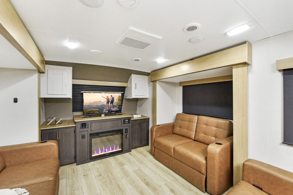 Modern RV living area with tan leather sofas, a TV above a built-in electric fireplace, light wood flooring, and white cabinetry. The room is brightly lit with ceiling lights and has large windows with black shades.