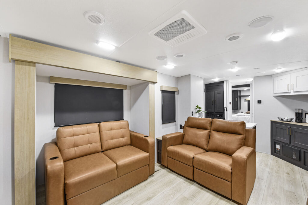 Modern RV interior with two tan leather couches facing each other, light wood flooring, white walls and ceiling, and built-in cabinets. Kitchen and bedroom areas visible in the background.
