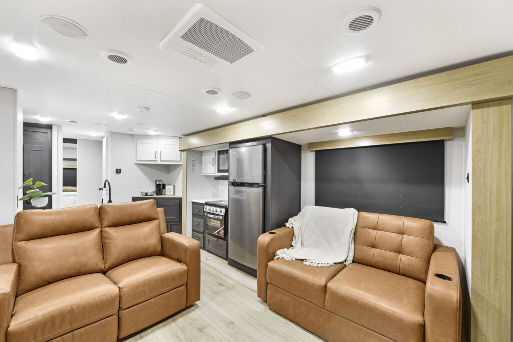 Modern RV interior with two tan leather sofas, a small kitchen with stainless steel appliances, light wood flooring, white walls, and recessed lighting. One sofa has a white blanket draped over the arm.