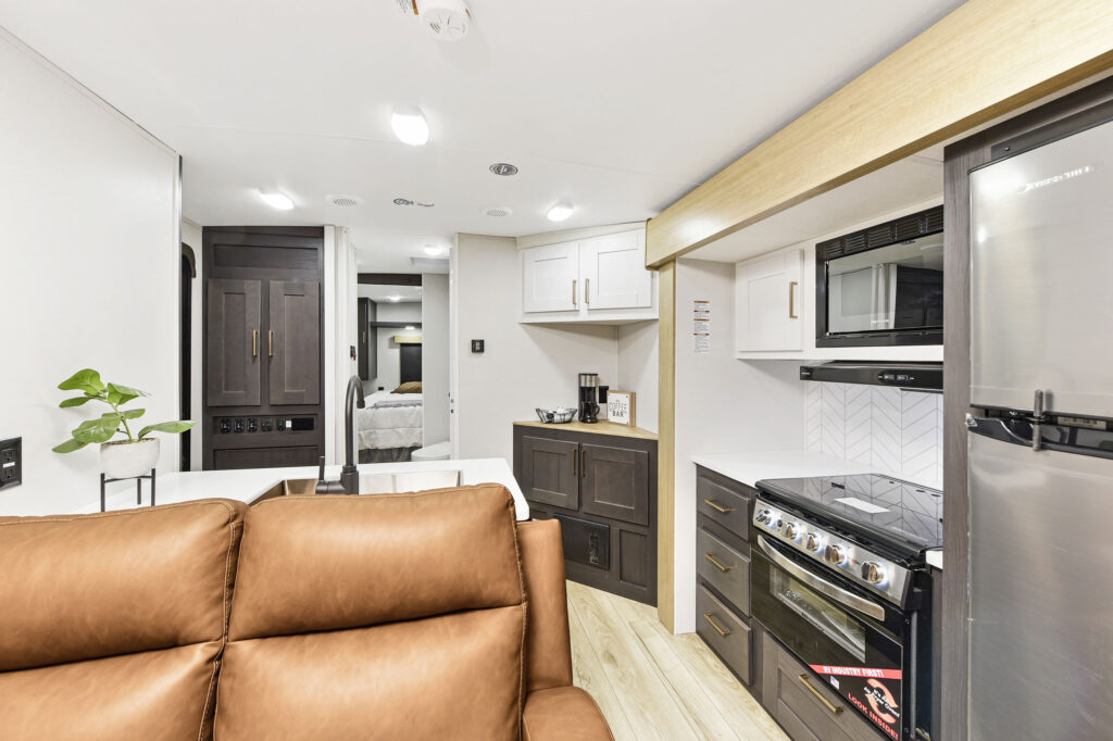 Modern RV interior featuring a brown leather sofa, compact kitchen with stove, microwave, fridge, white cabinets, dark lower cabinets, and a small potted plant; sleeping area visible in the background.